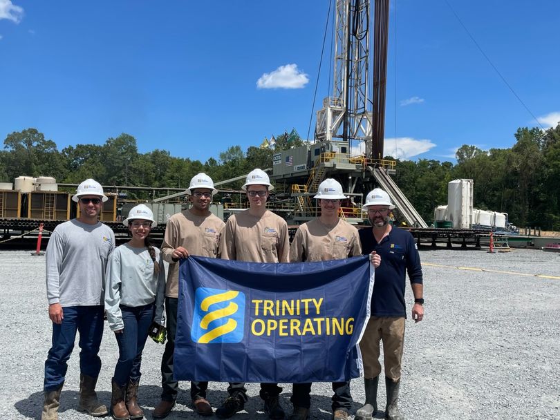 Six people hold a flag in front of energy extraction equipment. 