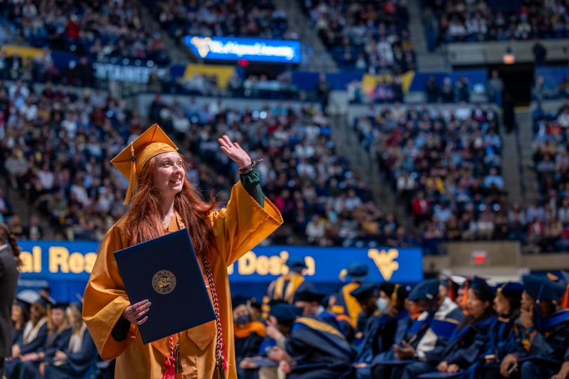 Female in gold cap and gown holding diploma and waving to crowd.