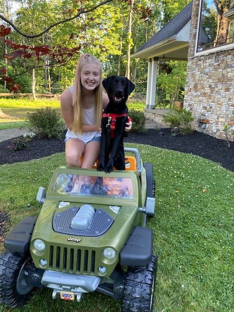 Photo of teenage girl sitting in toy car next to black dog.